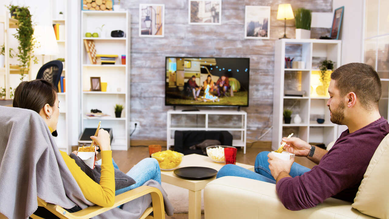 Casal assistindo a televisão enquanto comem macarrão 