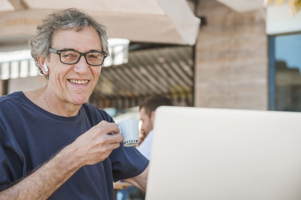 Idoso posa sorridente tomando café enquanto trabalha no notebook usando fones de ouvido bluetooth